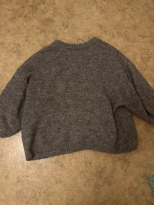 Grå stickad tröja med rund hals - En enkel och stilren grå stickad tröja med rund hals och är en trekvartärmad tröja. Tröjan har en avslappnad passform och ribbstickade muddar vid hals, ärmslut och nederkant. Perfekt för lager-på-lager och att hålla sig varm under kyliga dagar.