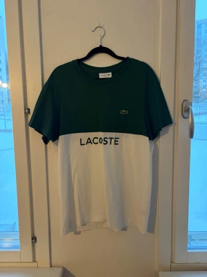 Grön och vit Lacoste t-shirt - Snygg t-shirt från Lacoste med grön ovandel och vit underdel. Klassisk logga på bröstet och stor Lacoste-text framtill. T-shirten har rund hals och korta ärmar, perfekt för en avslappnad stil.