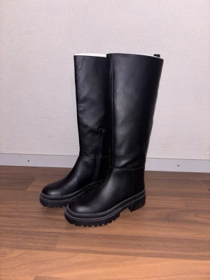 Svarta höga chunky stövlar från NA-KD - Säljer ett par svarta höga stövlar  med chunky sula och dragkedja på insidan från NA-KD. Stövlarna är i slätt konstläder och har en stilren, minimalistisk design. Perfekta för att lyfta din outfit med en edgy touch.