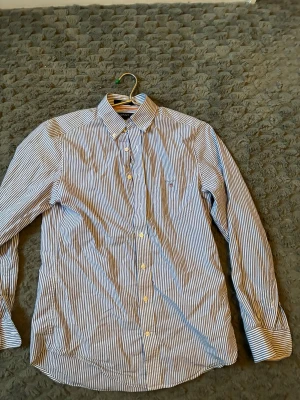 Blårandig skjorta från Gant - Klassisk skjorta från Gant med smala blå och vita ränder. Skjortan har knappar framtill, lång ärm och button-down krage. Liten broderad logga på bröstet. Perfekt för en stilren och tidlös look.