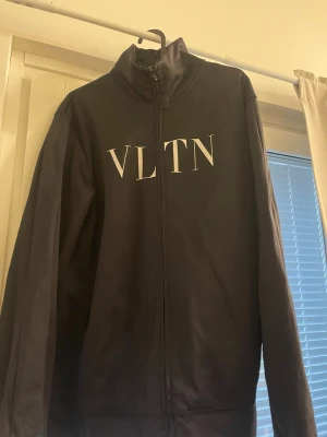 Valentino  - Svart tröja från Valentino med VLTN-tryck i vitt på bröstet. Tröjan har hög krage, hel dragkedja framtill och långa ärmar. Storlek är Medium i herrstorlek.