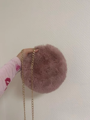 Rund fluffig axelväska i rosa fuskpäls - Supergullig rund axelväska i mjuk rosa fuskpäls med en snygg guldfärgad kedja som axelrem. Perfekt för att addera en söt och trendig touch till din outfit. Väskan är liten och smidig, perfekt för det viktigaste.