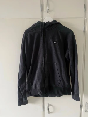 Svart zip hoodie från Nike - Säljer en svart hoodie från Nike med dragkedja framtill och två stora fickor. Tröjan har huva med dragsko och en liten Nike-logga på bröstet. Tillverkad i mjukt och bekvämt material, perfekt för chill dagar eller när du vill ha en sportig look.