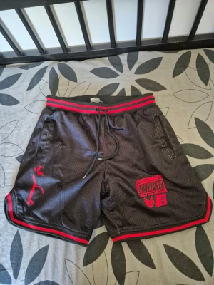 Askari Mesh Basketball Shorts – svart/röd – Storlek S - Svarta sportiga meshshorts från Askari Streetwear med röda tryck och detaljer. Shortsen har elastisk midja med snörning, röda ränder i midjan och längs benen samt tryck med siffror och logga både fram och bak. Perfekta för träning eller chill.