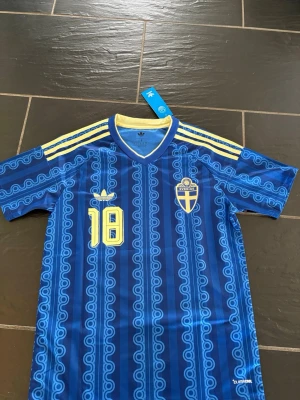 Blå Sverige fotbollströja Adidas S - Blå fotbollströja från Adidas med Sveriges landslagsemblem, gula detaljer och nummer 18 på bröstet. Tröjan har mönstrade blå ränder, gula axelband och v-ringad krage med gul insida. Tillverkad i lätt och ventilerande material (Climacool).