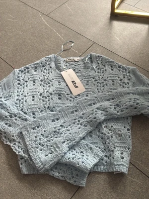 Ljusblå virkad tröja FB Sister - Söt ljusblå stickad tröja från FB Sister med virkat mönster och lång ärm. Tröjan har en avslappnad passform och är perfekt för lager-på-lager. Materialet är mjukt och luftigt, vilket gör den skön att bära under vår och sommar.