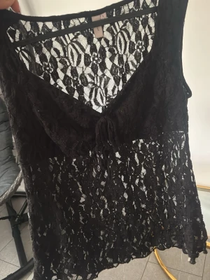 Svart spetslinne med knyt - Säljer ett svart, ärmlöst linne i genombruten spets med blommigt mönster. Toppen har v-ringning och en liten knytning framtill. Perfekt för dig som gillar en edgy och trendig stil.