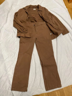 Brun byxdress från Kappahl - Snygg brun byxdress från Kappahl i storlek 38. Byxdressen har raka ben, stora fickor framtill och skjortkrage. Tillverkad i ett mjukt bomullsliknande material som känns bekvämt och avslappnat. Perfekt för dig som gillar en enkel men cool stil.