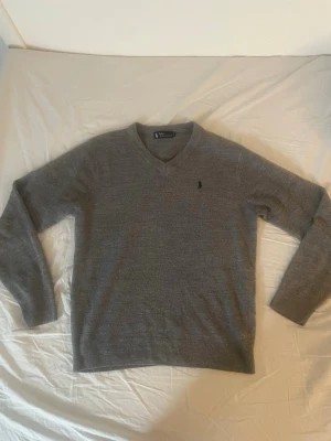 Grå stickad tröja Polo Ralph Lauren - Snygg grå stickad tröja från Polo Ralph Lauren med V-ringning och diskret logga broderad på bröstet. Tröjan har långa ärmar och ribbade muddar vid ärmslut och nederkant. Perfekt till lager på lager och enkel att matcha. Är i storlek XL men passar mer som L