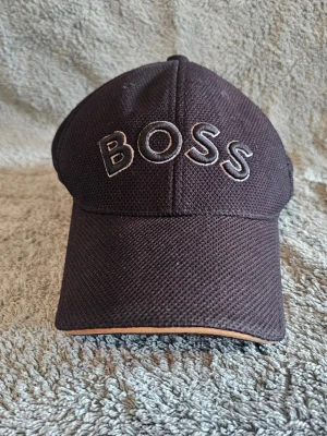Svart keps från BOSS - Snygg svart keps från BOSS med broderad logga framtill. Kepsen har böjd skärm, justerbart spänne bak och är tillverkad i ett ventilerande mesh-liknande material. Perfekt accessoar för en sportig och stilren look.