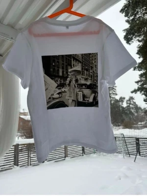 Vit t-shirt med retro fototryck - Vit t-shirt i bomull med ett stort svartvitt retro fototryck på ryggen som föreställer en stadsscen. Klassisk passform och rund halsringning. Perfekt för dig som gillar stilrena plagg med en twist.