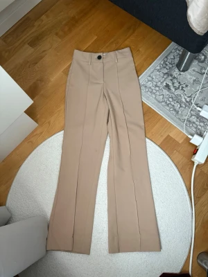 Beige kostymbyxor med pressveck - Snygga beige kostymbyxor med hög midja och vida ben. Byxorna har pressveck framtill, bälteshällor och stängs med knapp och dragkedja. Perfekta för en clean och stilren look. Materialet känns slätt och har lite struktur.