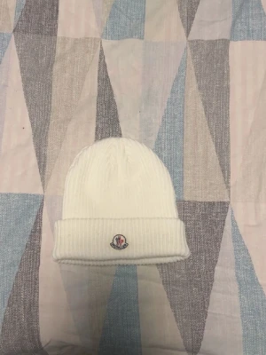 Vit ribbstickad mössa från Moncler - Snygg vit ribbstickad mössa från Moncler med uppvikt kant och klassisk Moncler-logga framtill. Tillverkad i mjukt material som håller dig varm och stilren under kalla dagar. Perfekt accessoar för att lyfta din vinterlook.