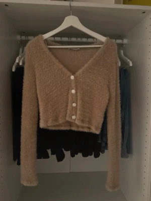 Beige fluffig kofta med blommiga diamant knappar - Supersöt beige fluffig kofta med v-ringning och långa ärmar. Koftan har fyra blommiga, glittriga knappar framtill och är croppad i modellen. Perfekt för dig som gillar mysiga och trendiga plagg med en gullig touch. Den är i ett jätte bra skick. De är en storlek L men den passar en storlek M/S med jag som är S kan ha på mig den och den sitter bra enligt mig.