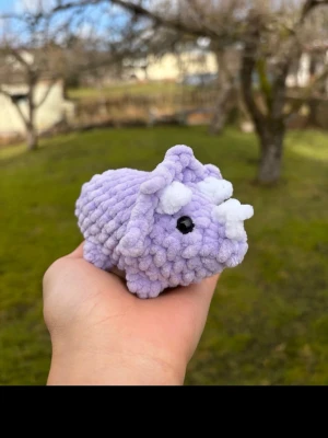 Lila virkad triceratops - Söt handvirkad triceratops i mjukt lila garn med vita horn och svarta ögon. Perfekt som dekoration eller gosedjur. Formen är rund och kompakt, och materialet är plyschliknande garn.