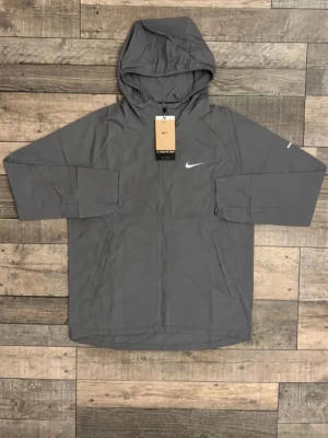 Grå Nike vindjacka med huva - Snygg grå vindjacka från Nike med huva och dragkedja framtill. Jackan har två sidofickor och en vit Nike-logga på bröstet. Tillverkad i lätt och smidigt material, perfekt för blåsiga dagar. Enkel och stilren design som passar till det mesta.