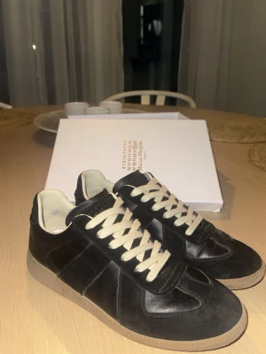Svarta Maison Margiela sneakers - Snygga svarta sneakers från Maison Margiela med beige sula och vita snören. Skorna har en klassisk låg siluett och är tillverkade i läder med detaljer i mocka. Perfekta för dig som gillar stilrena och trendiga sneakers. Box och allt orginalt medföljer.