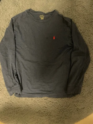 Mörkgrå långärmad tröja Polo Ralph Lauren - Storlek barn 14-16 160cm.Mörkgrå långärmad tröja från Polo Ralph Lauren med klassisk rund halsringning och röd broderad logga på bröstet. Tröjan är i mjuk bomull och har en avslappnad passform, perfekt för en stilren och enkel look.