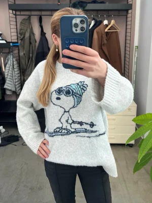 Ljusgrå stickad tröja med Snoopy - Mysig ljusgrå stickad tröja med en stor Snoopy-figur på framsidan. Tröjan har avslappnad passform, rund hals och ribbade muddar. Jättegosig och varm samt helt ny.