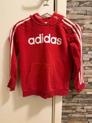 Röd adidas hoodie med vita ränder - Snygg röd hoodie från adidas med klassiska vita ränder längs ärmarna och stor vit logga på bröstet. Huva med adidas-tryck och stor magficka framtill. Perfekt för en sportig och avslappnad stil. Tillverkad i mjukt bomullsmaterial.
