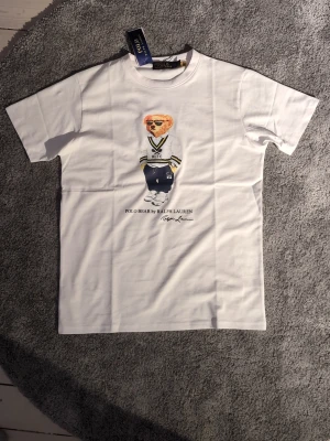 Vit t-shirt Polo Ralph Lauren - Vit t-shirt från Polo Ralph Lauren med ikoniskt björntryck på bröstet. Klassisk passform med korta ärmar och rund halsringning. Den är i nytt skick utan prislappen