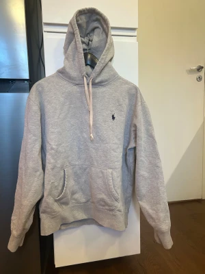 Grå hoodie från Polo Ralph Lauren - Snygg grå hoodie från Polo Ralph Lauren med klassisk huva och dragsko. Har en broderad logga på bröstet och en stor magficka. Tillverkad i mjuk bomullsblandning, perfekt för en chill och avslappnad stil.