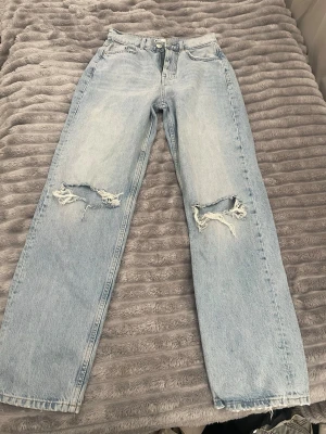 Ljusblå ripped jeans med raka ben - Säljer ett par ljusblå jeans med raka ben och slitna hål över knäna. Jeansen har hög midja, klassisk femficksdesign och råa kanter nertill. Perfekta för en avslappnad och trendig look.