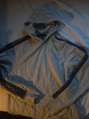 Grå hoodie från GANT med logga - Snygg grå hoodie från GANT med svarta band längs ärmarna där GANT-loggan är tryckt i vitt. Klassisk huva och ribbade muddar vid ärmslut och nederkant. Tillverkad i mjukt bomullsmaterial för skön känsla. Perfekt för en avslappnad och trendig look.