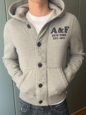 Grå hoodie från Abercrombie & Fitch - Säljer en Abercrombie & Fitch hoodie i fint skick. Inga defekter, knappt använd och fortfarande väldigt fräsch. Perfekt som en stilren och bekväm basic till vardags.  Modellen på bilden är 187 cm lång och väger 80 kg för referens på passform.  Snabb leverans 🚚