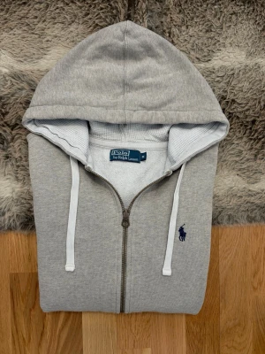 Ljusgrå Polo Ralph Lauren Hoodie - Hej! 👋 Säljer en klassisk ljusgrå Polo Ralph Lauren hoodie i storlek M med mörkblå pony-logga. 🐎 Tröjan är i mycket gott skick i övrigt, men jag vill vara helt transparent med att det finns en pytteliten slitning/hål precis under bröstet (se närbild). Det är inget som stör jättemycket när man bär den, men priset är satt därefter! ✨ 100% äkta med tvättlappar kvar. Ingen QR-kod. Perfekt som en skön vardagströja eller för dig som vill ha ett premiumplagg till ett riktigt bra pris. Fraktar snabbt! 🚛