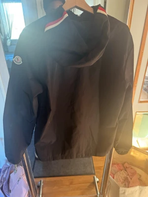 Svart Moncler vindjacka med huva - Svart vindjacka från Moncler med huva och snygga röda och vita detaljer på luvan. Jackan har Moncler-logga på vänster ärm och en stilren, sportig look. Perfekt för blåsiga dagar och enkel att matcha med olika outfits.