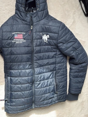 Mörkblå pufferjacka från Vinson Polo Club - Snygg mörkblå pufferjacka från Vinson Polo Club med broderad logga och amerikansk flagga på bröstet. Jackan har huva med dragsko, två fickor med dragkedja och ribbade muddar vid ärmsluten. Perfekt för kalla dagar.