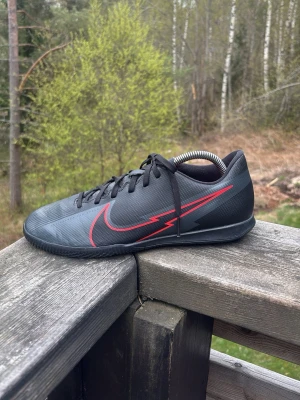Nike svart-röda fotbollsskor - Säljer ett par svarta Nike fotbollsskor med röd blixtdesign och klassisk Swoosh-logga på sidan. Skorna har låg profil, snörning och tunn yttersula som passar perfekt för spel på inomhusplan som till exempel innebandy eller futsal. Snygg och sportig look för dig som vill sticka ut på planen.