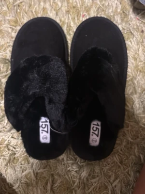 Svarta fluffiga tofflor från 157 - Mysiga svarta tofflor från 157 med fluffig fuskpäls upptill och mjuk innersula. Yttersulan är i slitstark gummi och ger bra grepp. Perfekta för att hålla fötterna varma hemma. Klassisk slip-in modell som är enkel att ta på.