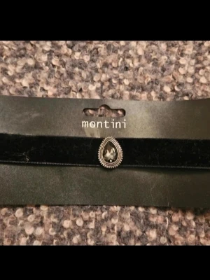 Svart choker med droppformad sten Montini - Snygg svart choker från Montini med en droppformad, mörkgrå sten i mitten. Halsbandet är gjort av sammet och har en dekorativ metallram runt stenen. Perfekt accessoar för att ge din outfit en cool och elegant touch.
