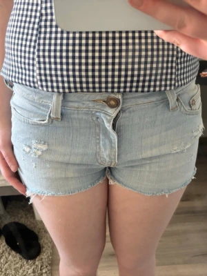 Ljusblå jeansshorts med slitningar - Säljer ett par ljusblå jeansshorts med råa kanter och slitna detaljer framtill. Klassisk femficksmodell med knapp och dragkedja. Perfekta för varma dagar och enkel att matcha med allt.