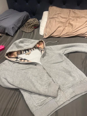 Burberry kofta - Snygg grå hoodie från Burberry med dragkedja och klassiskt rutigt foder i huvan. Hoodien har fickor framtill och broderad logga på bröstet. Perfekt för en stilren och avslappnad look.