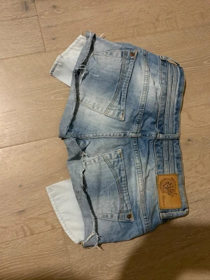 Blå jeansshorts med rå kant - Snygga blå jeansshorts från Fishbone med rå, fransig kant och dubbla knappar framtill. Klassisk femficksmodell och låg midja. Perfekta för varma dagar och enkel att matcha med olika toppar.Waist 35cm Rise 18cm Storlek 26