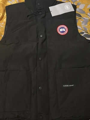 Svart dunväst från Canada Goose - Passar storlek M eller stor L. Svart dunväst från Canada Goose med klassisk logotyp på bröstet och två stora fickor framtill. Västen har hög krage, tryckknappar och diskret varumärkesmärke på ena fickan. Perfekt för lager-på-lager och kyliga dagar.