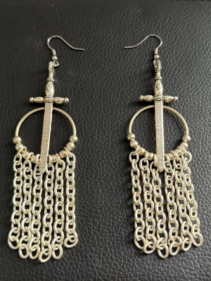 Silvriga örhängen med svärd och kedjor - Unika örhängen i silverfärgad metall med svärdformad detalj och flera hängande kedjor. Designen är medeltid inspirerad och har en rund ring med coola detaljer på svärdet. Nypris : 700kr 