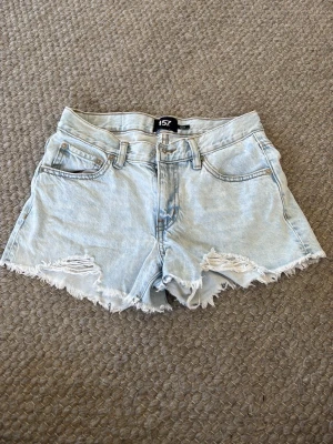 Ljusblå denimshorts från 157 - Säljer ett par ljusblå denimshorts från 157 med råa, fransiga kanter och slitningar framtill. Klassisk femficksdesign och hög midja. Perfekta för varma dagar och en avslappnad look.