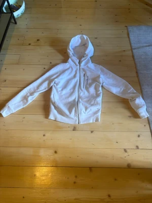 Vit hoodie från Sail Racing - Säljer en vit hoodie från Sail Racing med hel dragkedja och huva. Den har en snygg bröstficka med dragkedja och logotyp på vänster sida. Materialet är mjukt och bekvämt, perfekt för chill dagar. Passformen är slim och ärmarna har ribbade muddar.