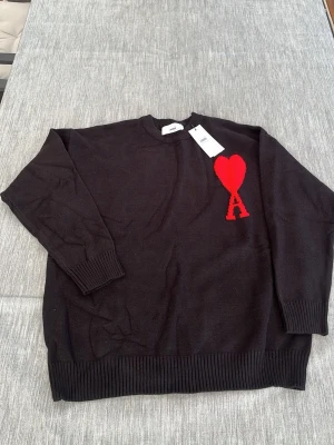 Svart stickad tröja från Ami Paris - Svart stickad tröja från Ami Paris med rund halsringning och ribbade muddar. På bröstet finns ett stort rött hjärta med bokstaven A, vilket är varumärkets ikoniska logga. Tröjan har en avslappnad passform och är perfekt för lager-på-lager-stil.
