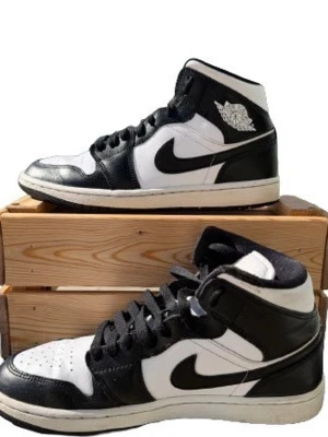 Svarta och vita Air Jordan 1 High - Snygga Air Jordan 1 High sneakers i svart och vitt läder med klassisk Nike Swoosh på sidorna och Air Jordan-logga vid ankeln. Skorna har svart snörning, perforerad tåbox och robust gummisula. Perfekta för dig som gillar streetwear och ikonisk design.