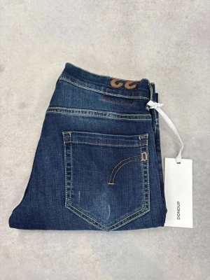 Mörkblå slim fit jeans från Dondup - Snygga mörkblå jeans från Dondup med slim fit passform och klassiska fem fickor. Jeansen har diskreta slitningar och kontrastsömmar, samt en läderpatch med logga bak i midjan. Perfekta för en trendig och avslappnad look.