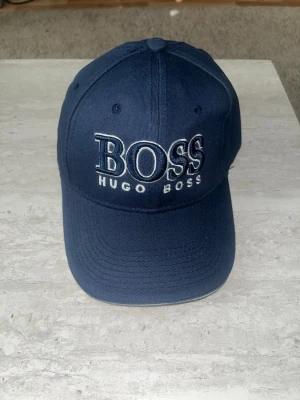 Mörkblå keps från Hugo Boss - Snygg mörkblå keps från Hugo Boss med stor broderad BOSS-logga framtill. Klassisk böjd skärm och justerbar rem baktill. Kepsen är tillverkad i bomull och har ventilerande hål upptill för extra komfort.