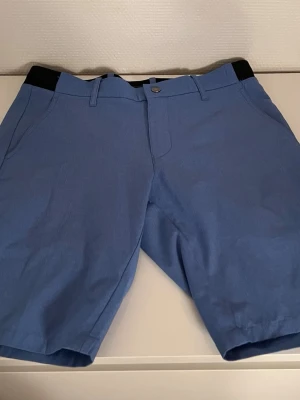 Blå shorts från Abacus W28 - Blå shorts från Abacus med klassisk passform och bälteshällor. Shortsen har svarta detaljer i midjan, knapp och dragkedja framtill samt sidofickor. Tillverkade i ett lätt och bekvämt material som passar perfekt för sommaren.