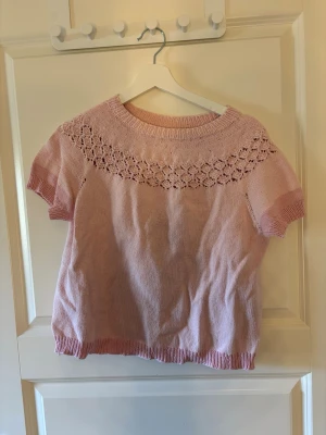 Ljusrosa stickad topp med spetsmönster - Söt ljusrosa stickad topp med korta ärmar och vackert spetsmönster runt halsringningen. Tröjan har ribbstickade muddar vid ärmslut och nederkant. Perfekt för dig som gillar en feminin och mjuk stil.