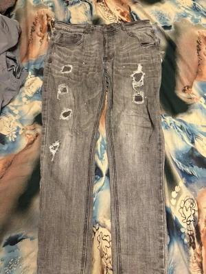 Grå slitna jeans från Alessandro Zavetti - Säljer ett par grå jeans från Alessandro Zavetti med flera slitningar och hål på framsidan. Jeansen har klassisk femficksmodell, bälteshällor och dragkedja. Perfekta för en trendig och avslappnad streetstil.