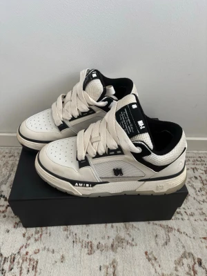 AMIRI vita och svarta sneakers - Snygga sneakers från AMIRI i vitt och svart läder med breda vita skosnören och perforerad tå. Skorna har svart innerfoder, detaljer i mesh och tydlig logga på sidan. Perfekta för dig som gillar streetwear och exklusiva märken.